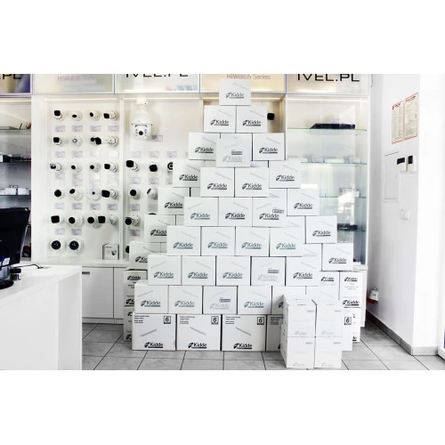 A stack of white boxes featuring the Kidde logo is displayed in a pyramid formation in a showroom. The background showcases various security and surveillance products on shelves.