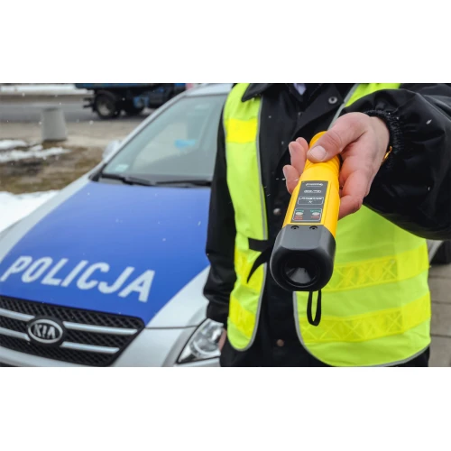 A person in a reflective vest holds a yellow breathalyzer device with a digital display, designed for quick and efficient alcohol testing. The background features a police car, indicating its use in law enforcement scenarios.