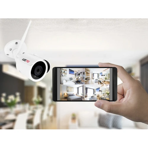 A wireless network camera is mounted on a ceiling, featuring a sleek white design with an antenna for enhanced connectivity. A hand holds a smartphone displaying a multi-view interface of various rooms, showcasing the camera's real-time monitoring capabilities.