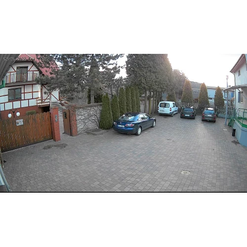 The image shows a driveway with parked cars and a brick wall, captured by a wireless network camera. The scene includes trees and a residential building, demonstrating the camera's outdoor surveillance capabilities.