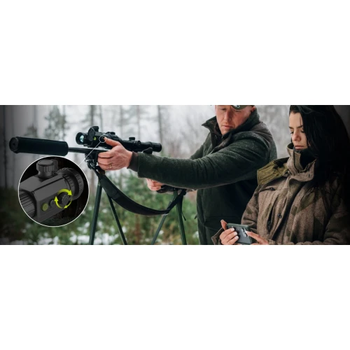 A man is adjusting a thermal imaging monocular mounted on a rifle, while a woman holds a control device. The scene is set outdoors in a forested area, highlighting the product's use in natural environments.