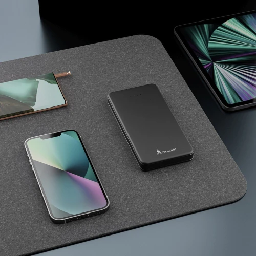 A sleek black power bank with the Extralink logo is placed on a dark textured surface, surrounded by modern electronic devices. The power bank features a minimalist design, complementing the aesthetic of the nearby smartphone and tablet.