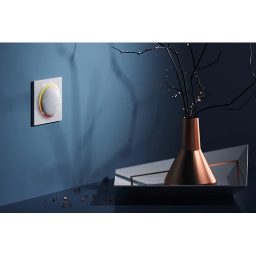 A sleek, modern smart light switch with a circular design and subtle LED ring is mounted on a stylish blue wall. It is accompanied by a decorative copper vase and a geometric tray, creating an elegant and contemporary setting.