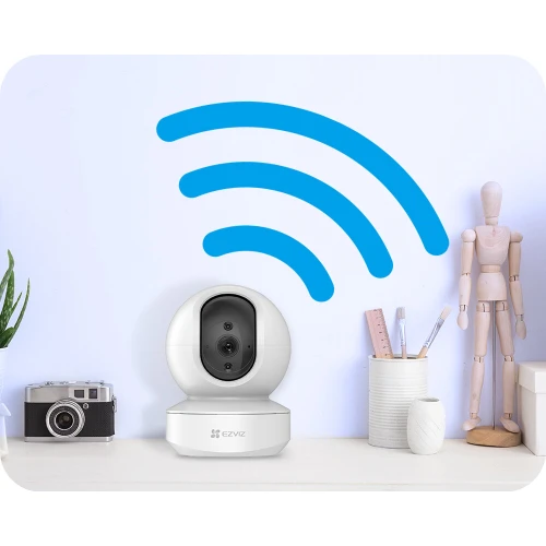 A sleek, white EZVIZ TY1 Full HD IP camera is positioned on a desk, surrounded by stationery and a wooden mannequin. A blue Wi-Fi symbol is displayed on the wall behind, indicating wireless connectivity.