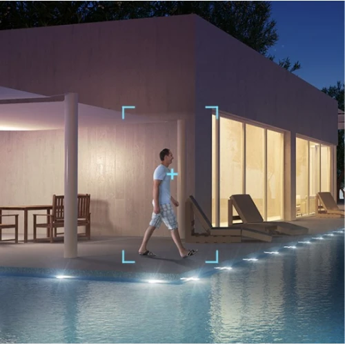 A modern outdoor security camera is mounted on a building, overlooking a pool area with lounge chairs and ambient lighting. The camera features motion detection, highlighted by a digital overlay tracking a person walking by.