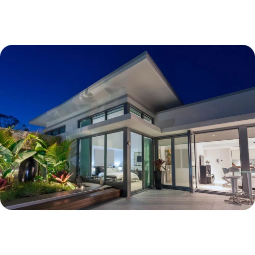 Modern, minimalist house with large glass windows and doors, featuring a sleek, angular roof design. The exterior is complemented by lush greenery and contemporary outdoor lighting, creating a welcoming ambiance.