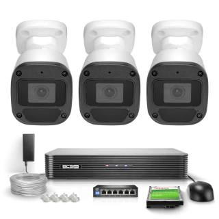 The image shows a BCS Basic 5M IP surveillance system, featuring three white security cameras and a central recording unit. Included are cables, connectors, and a power adapter for comprehensive monitoring setup.