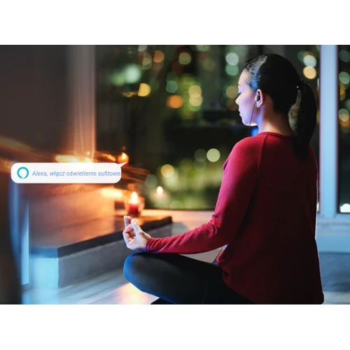 A woman sits in a meditative pose in a dimly lit room, surrounded by candles, while using a voice command to control the lighting. The scene highlights the convenience and ambiance created by smart home technology.