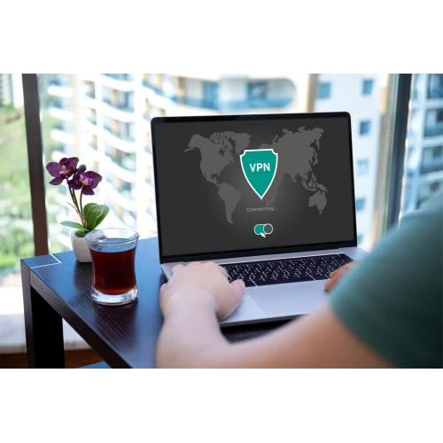 A person is using a laptop displaying a VPN connection screen, set on a table next to a glass of juice and a small potted plant. The background shows a view of tall buildings through a window.