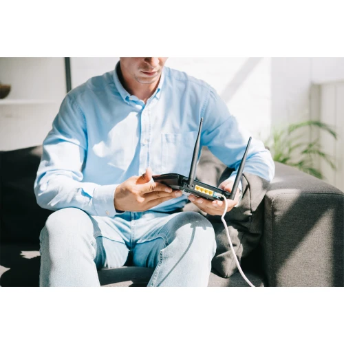 A person is sitting on a couch holding a black router with two antennas and several Ethernet ports visible. The router is connected to a white cable, suggesting it is ready for setup or use.
